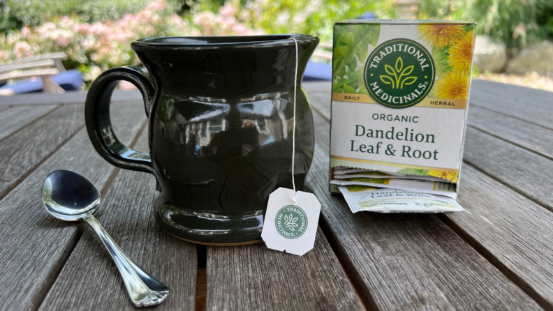 A mug with a tea bag string sit on an outdoor table between a spoon and a box of dandelion leaf and root tea.