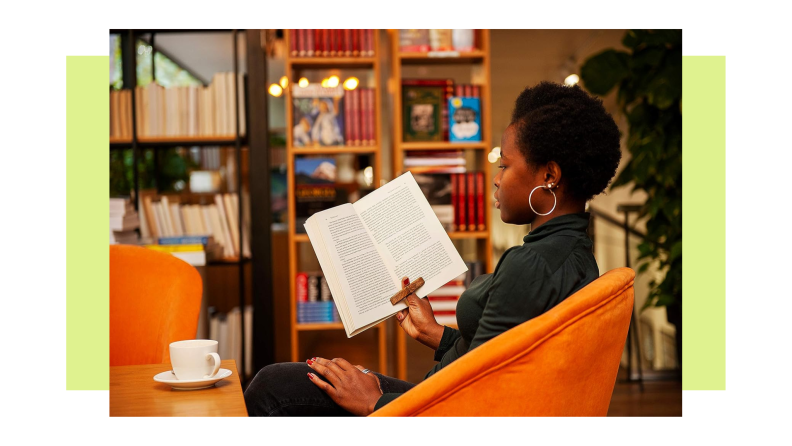 An African American woman sits in a chair, using the wooden Tilisma book holder to read a book. The holder is inserted into the thumb and features platforms on the left and right that hold open the pages.