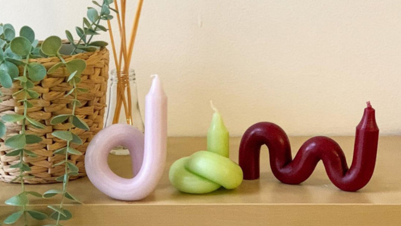 One pink twisted candle, one knotted green candle, and one bendy maroon candle next to each other on a table next to a plant