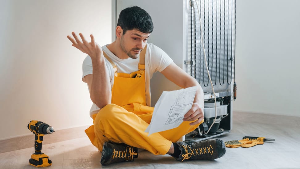 A man sits on the ground near a fridge, near a drill and some manuals. He's reading the fridge's warranty and putting up one hand in confusion at what he's reading.