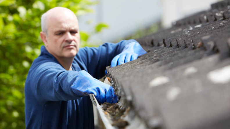 Person on ladder cleaning leaves out of dirty gutter outdoors.