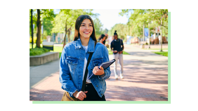 A smiling young student holding some textbooks wearing a messenger bag walking around a college campus.