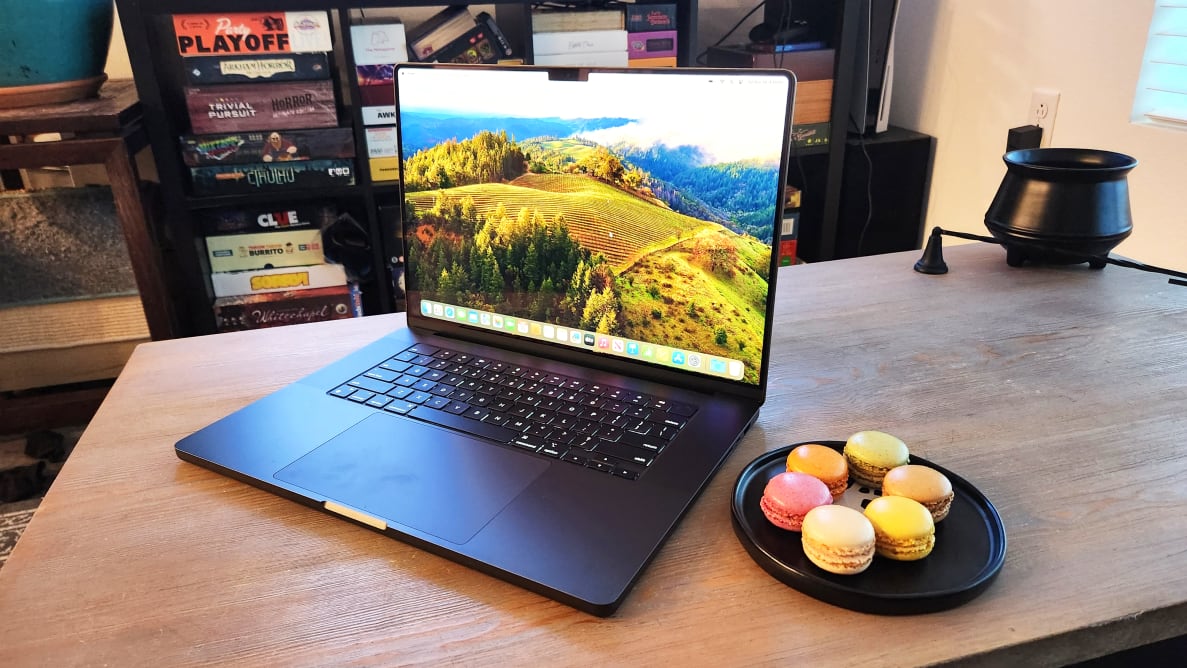 An open and powered on laptop on top of a wooden surface next to a black plate containing macaroons.