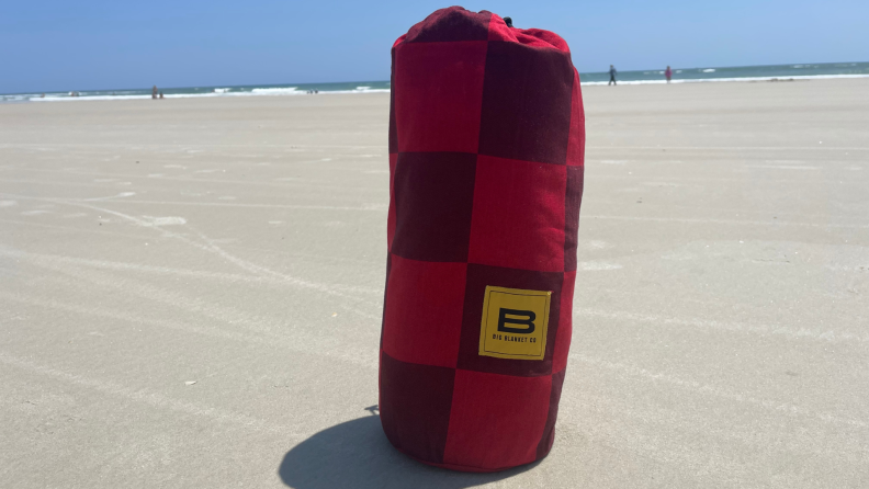 Red and burgundy checkered carrier on top of sand outdoors with Big Beachy Blanket inside.