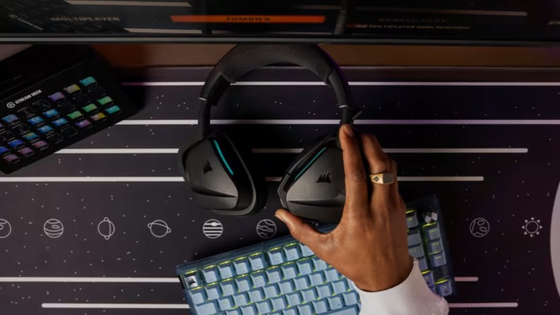 A hand reaches for a black Corsair headset resting on a space-themed gaming desk setup.
