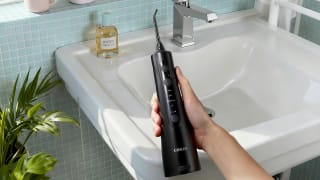 Hand holding a black COSLUS water flosser over a sink next to a potted plant