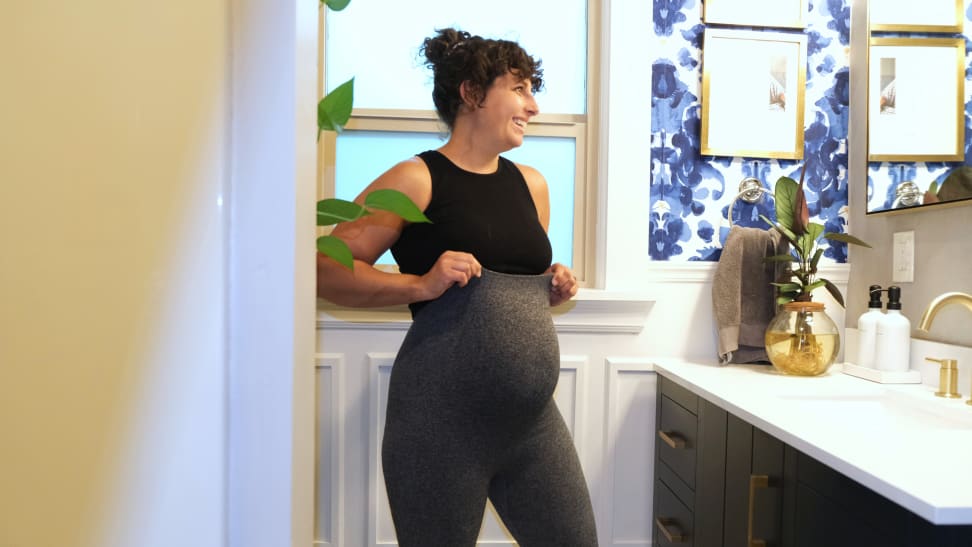 A woman standing in a bathroom pulling up gray leggings over her pregnant belly.