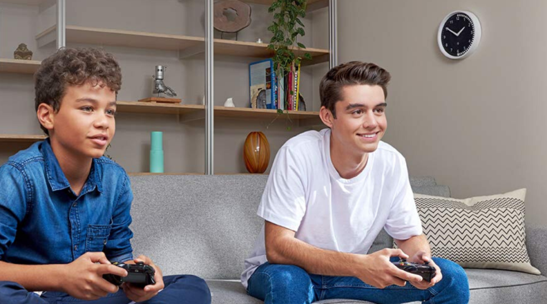 Two kids sit on the couch and play video games with the Echo Wall Clock displayed on the wall.