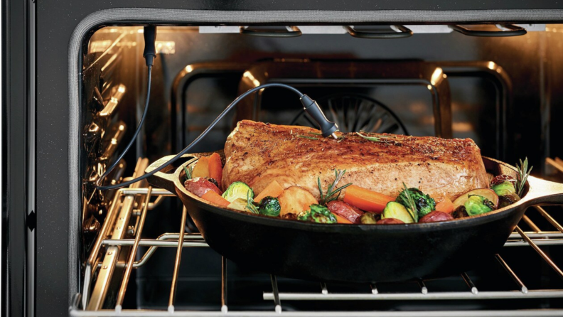 A pot of roasted pork with vegetables is in an oven. A meat thermometer is probed into the meat.