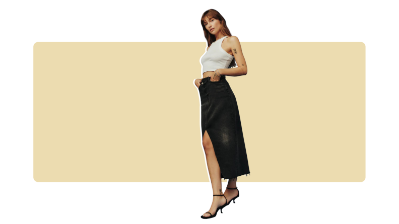 A model wearing a midi-length black denim skirt with a front slit and frayed hem. She also has on a white tank top.
