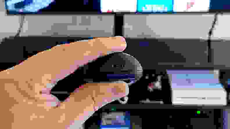 A hand holds the black, rounded remote in front of a TV console with a microUSB port showing.