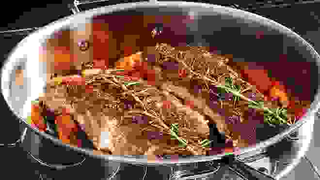 An All-Clad pan on a stove containing ribs on a bed of carrots and topped with rosemary.