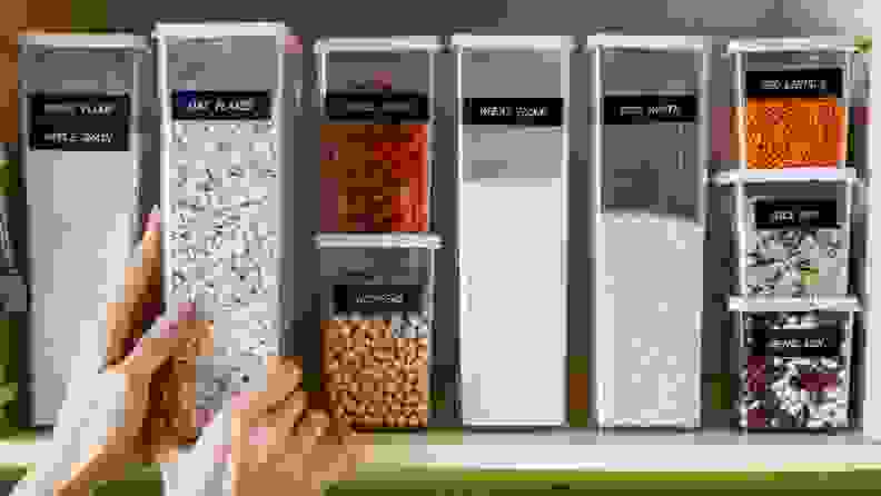 Food bins neatly organized and named in a pantry, with a hand reaching out for one of them.
