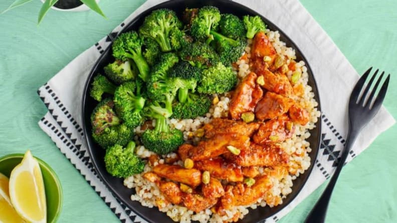Broccoli and harissa apricot chicken over couscous with pistachios on a black plate with a fork and slice of lemon.