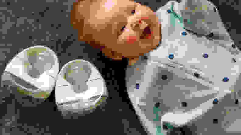 A smiling baby lies on a blanket beside a pair of breast pumps.