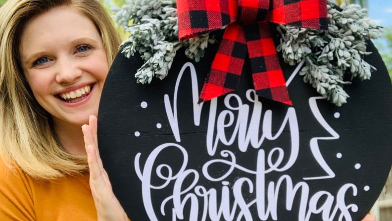 Woman holding a round wooden door hanger with a red plaid bow and artificial evergreens reading "Merry Christmas"