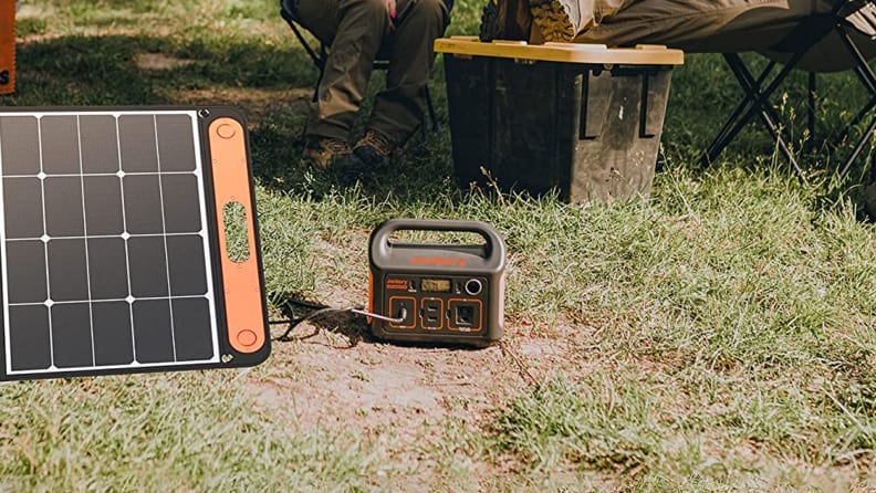 A power station sits on the ground in the woods, getting recharged by a nearby solar panel. In the background you can see two campers relaxing.
