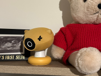 A yellow baby monitor sits on a shelf next to a brown teddy bear wearing a red sweater