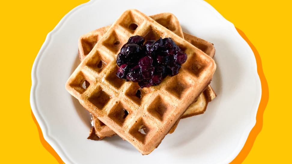 Two waffles with blueberry compote on top sitting on a white plate on a white surface.