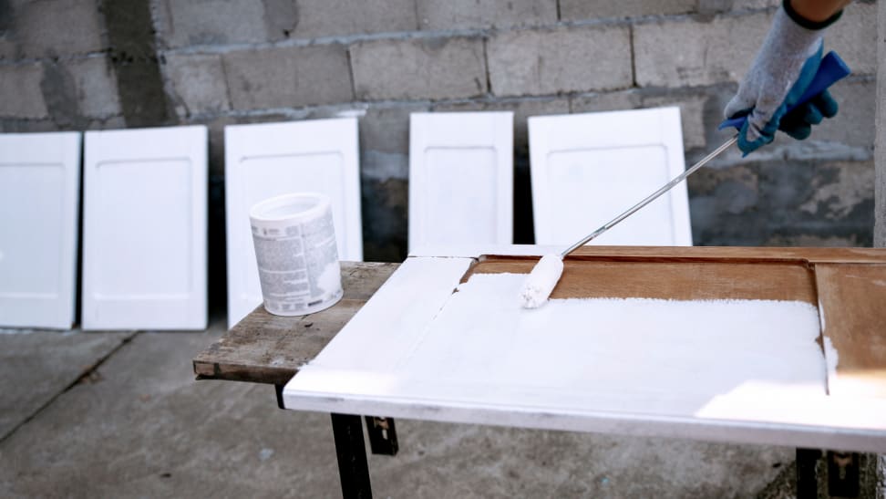 Gloved hand using paint roller to paint cabinets white outdoors.