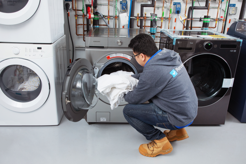 Person crouching down to place white fabrics inside of washer machine drum indoors.