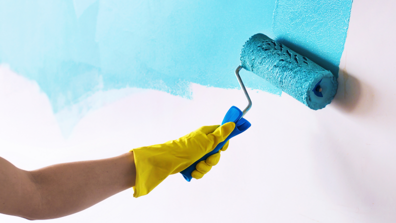 Hand wearing yellow rubber glove using paint roller to paint wall blue.