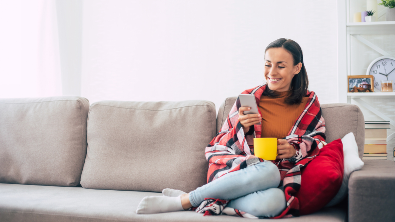 Person siting on couch under comfy blanket while looking at smartphone in living room.