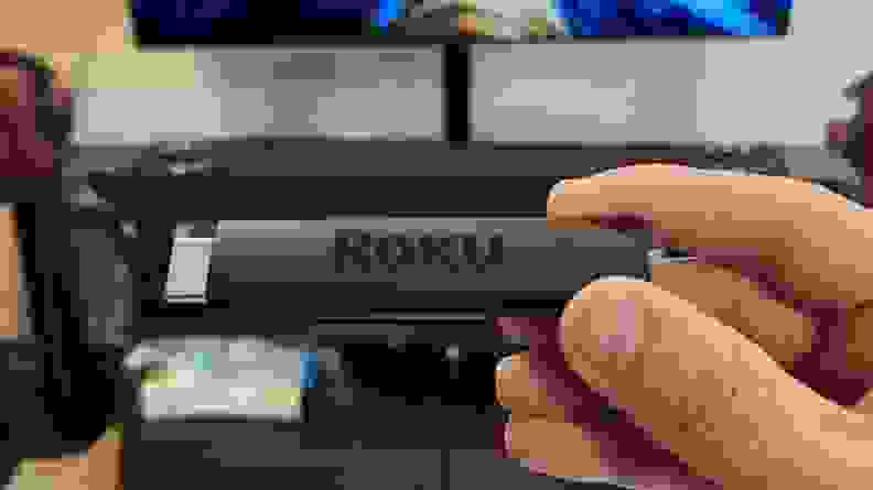 The matte black, candy-bar sized Roku Streaming stick is held with its port showing an OLED TV.