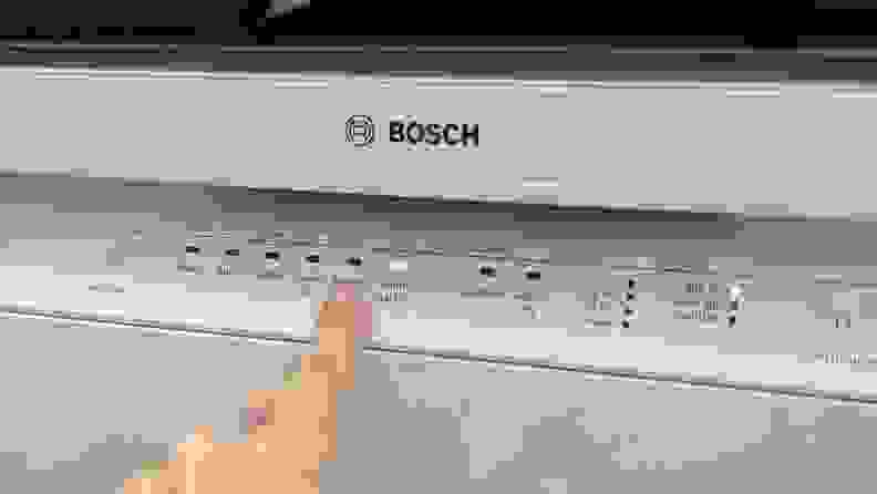A finger points to the "Favorite" cycle button on the front-facing control panel of the Bosch 100 Series SHE3AEM2N dishwasher.