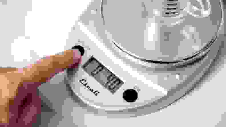 A hand presses a button on the Escali digital kitchen scale.