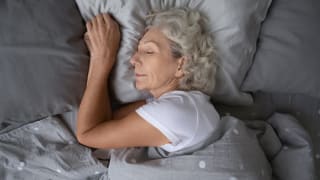 Older, white woman sleeping peacefully alone with grey bedding.