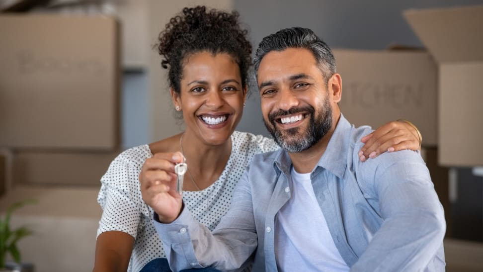 New homeowners hold up a key to their new house with cardboard moving boxes in background