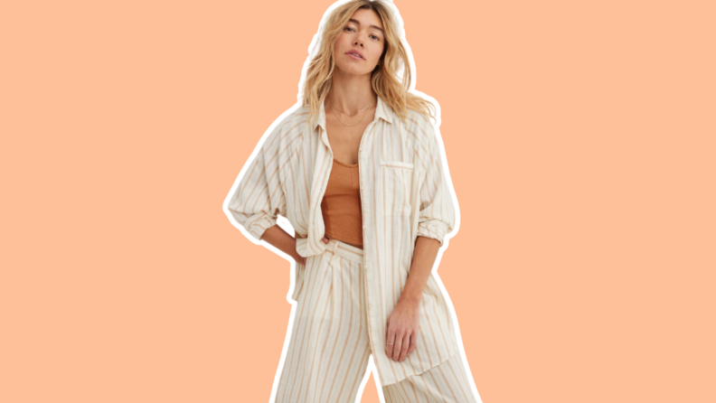 A model posing with their hand on hip while wearing a cream striped linen shirt and matching pants on an orange background.