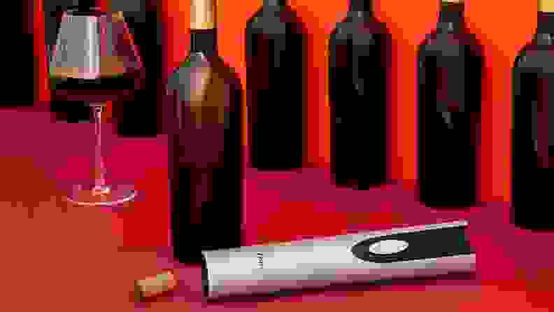 A cordless electric wine opener lays on a red counter beside an open bottle of wine and a half full wine glass.