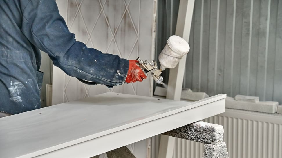 A person in protective clothing spraying white paint from a paint gun onto a piece of wood